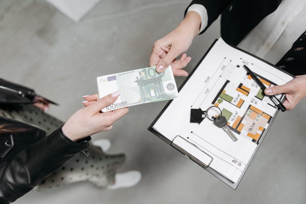 Hand passing a euro banknote over house keys and a floor plan on a desk
