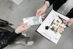 Hand passing a euro banknote over house keys and a floor plan on a desk